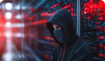 Person in a hoodie and face covering in front of illuminated servers with red and blue lights.