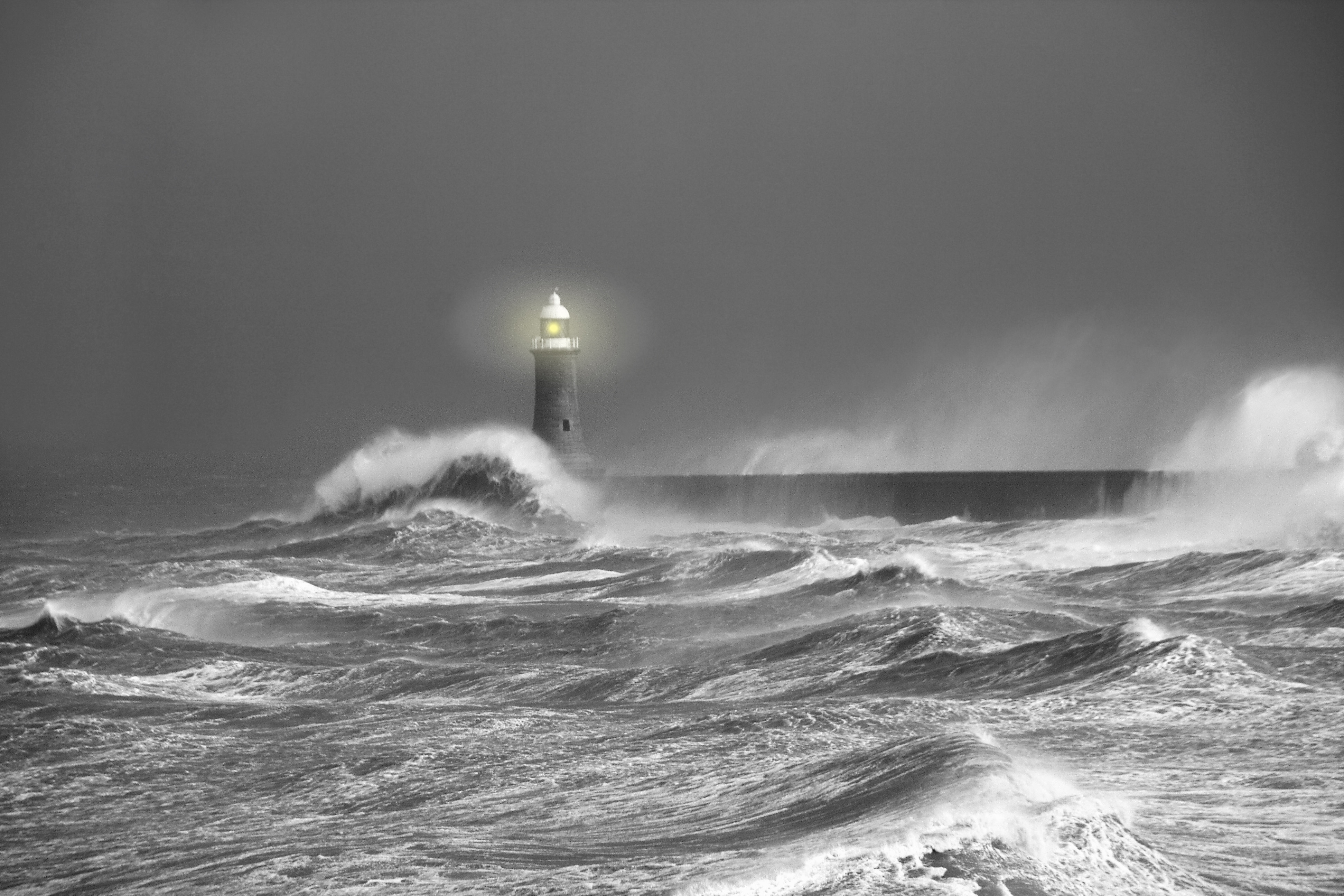 black and white photo of a raging ocean with waves pounding a lit lighthouse